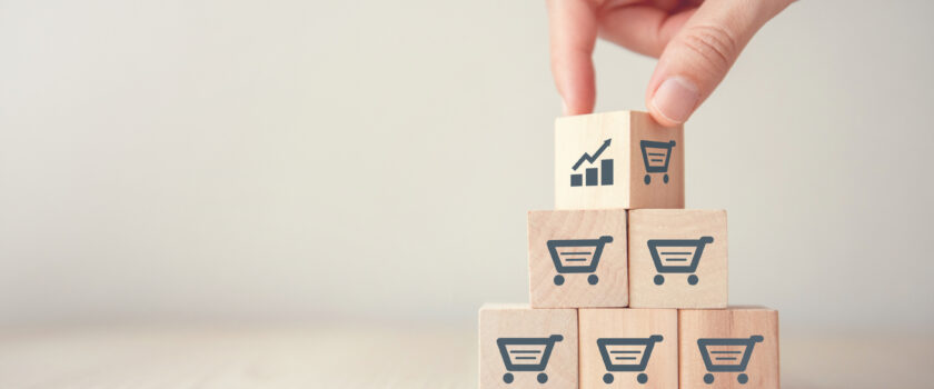A hand placing a wooden block with a growth graph icon atop a stack of blocks with shopping cart icons, symbolizing the strategic growth in e-commerce marketing.
